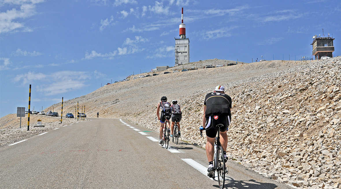 Three cyclists riding up a steep mountain road on a summer’s day, towards the summit which is marked by a red and white spire